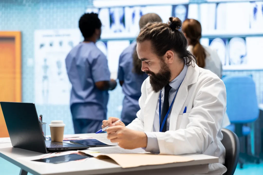 Un medico che controlla una cartella medica. Nello sfondo un altro medico e degli infermieri controllano dei raggi.