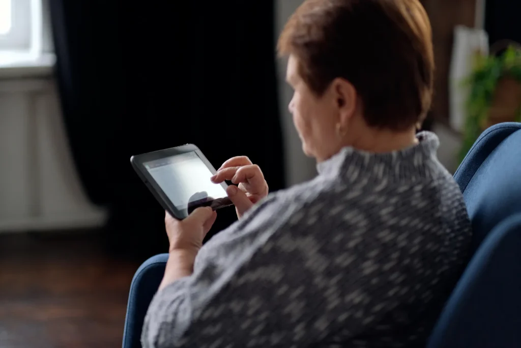 Donna anziana inquadrata di spalle: è seduta su una poltrona e utilizza un tablet con entrambe le mani.