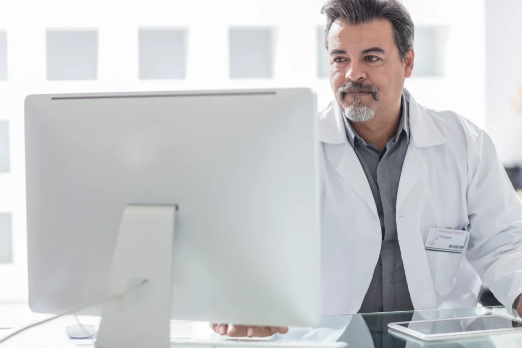 Medico in camice bianco seduto alla scrivania mentre consulta il monitor di un computer in un ambiente clinico luminoso.